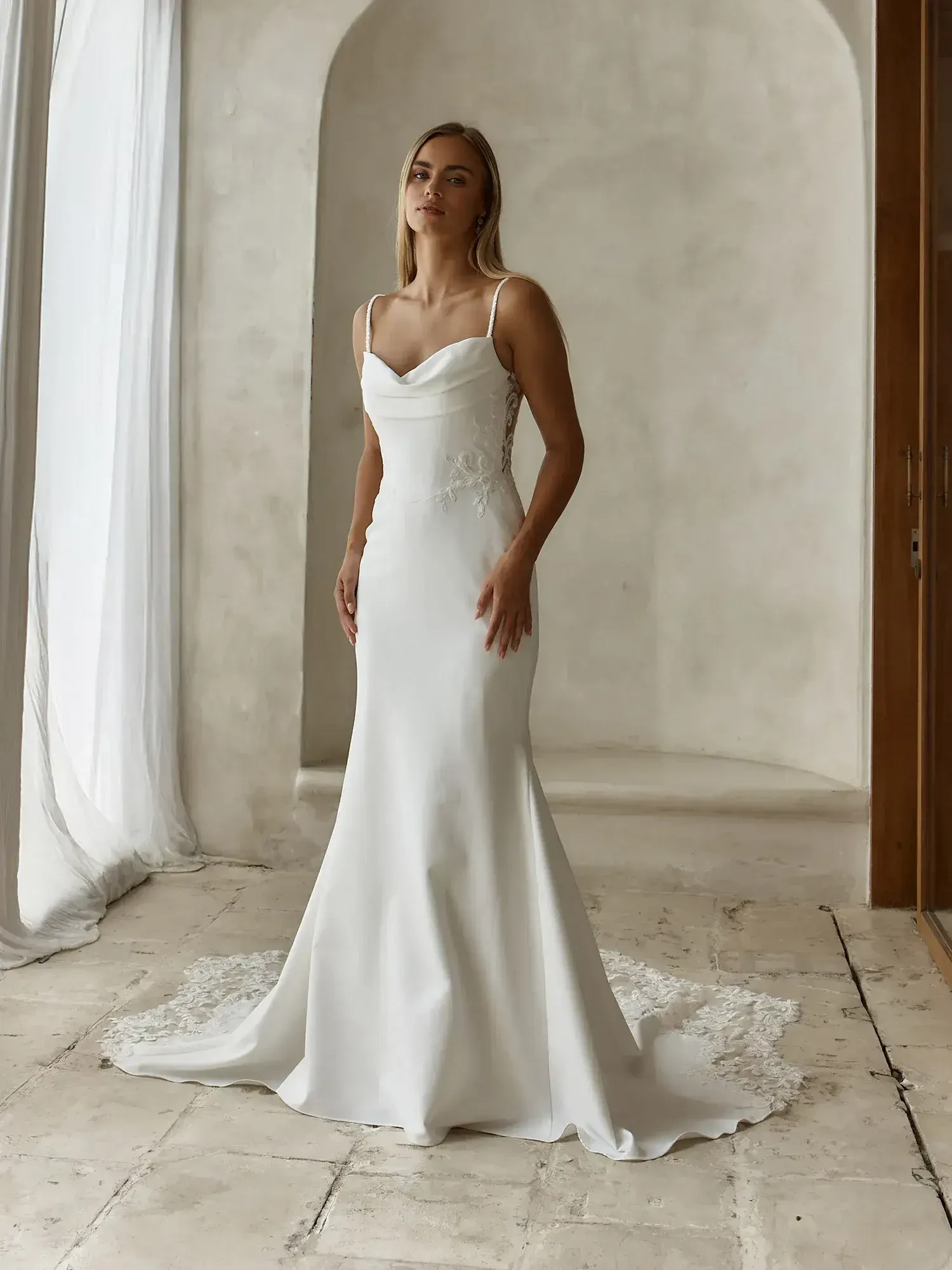 A woman wearing a fitted, elegant white wedding dress with thin straps, standing in a bright, softly lit room.