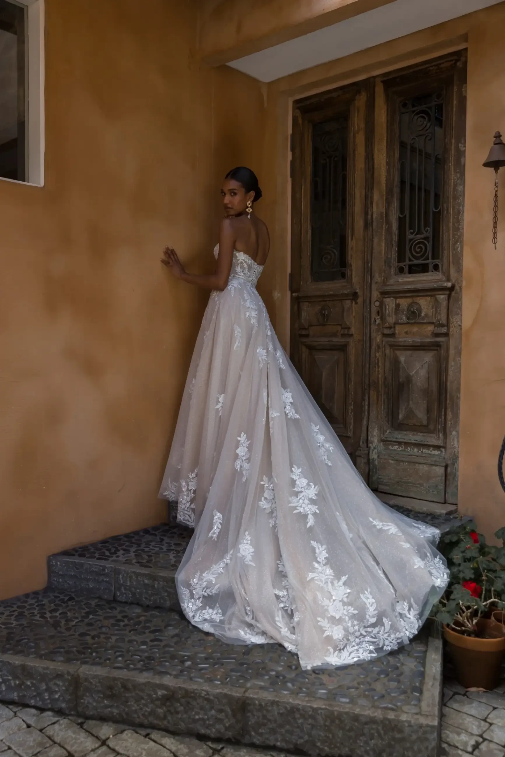 A woman in an elegant, floral lace wedding gown stands on stone steps by an aged wooden door, conveying a serene and timeless ambiance.