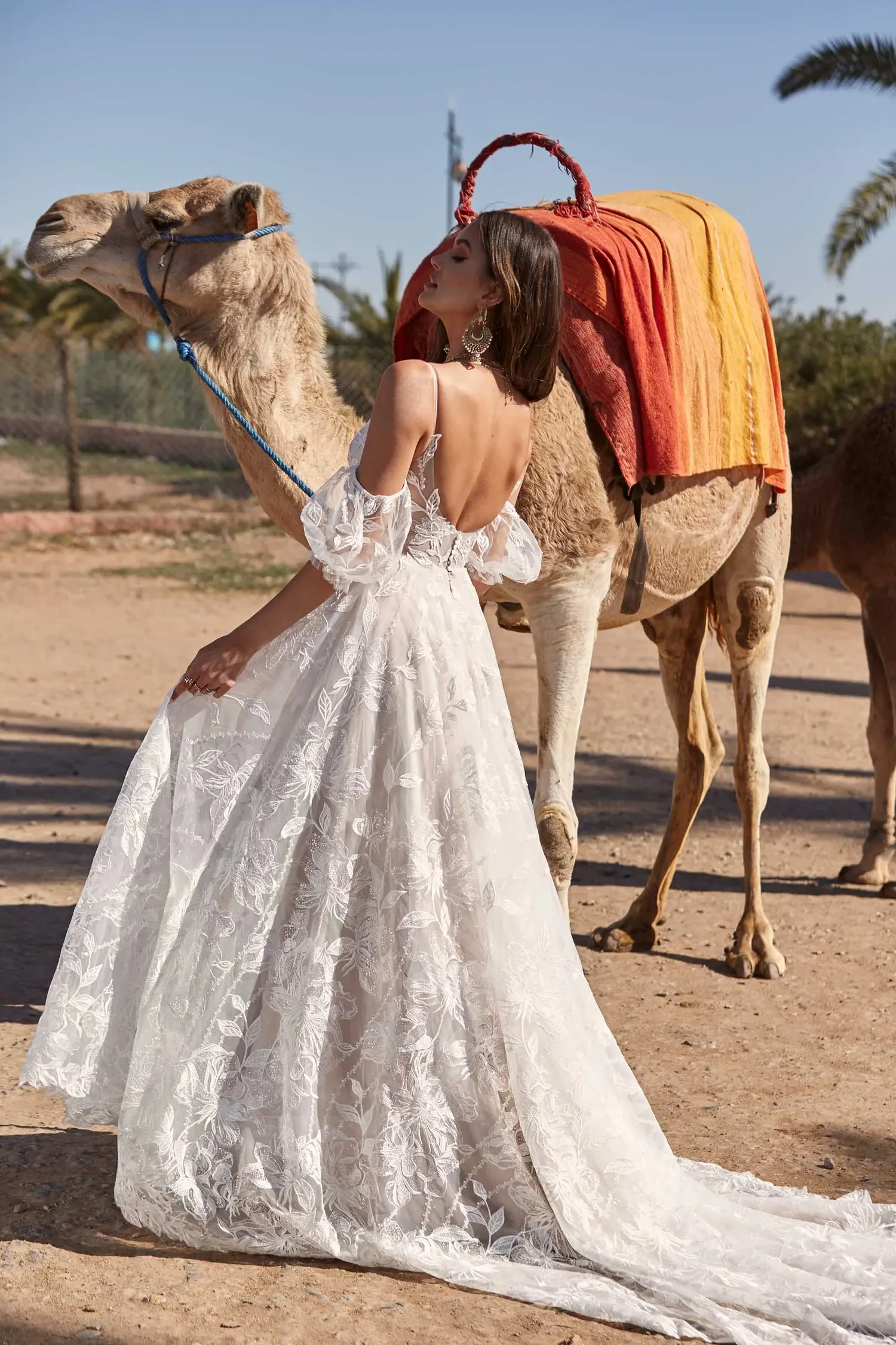 A woman in a flowing white lace gown stands beside a camel with a colorful blanket saddle, under a clear blue sky, conveying elegance and adventure.