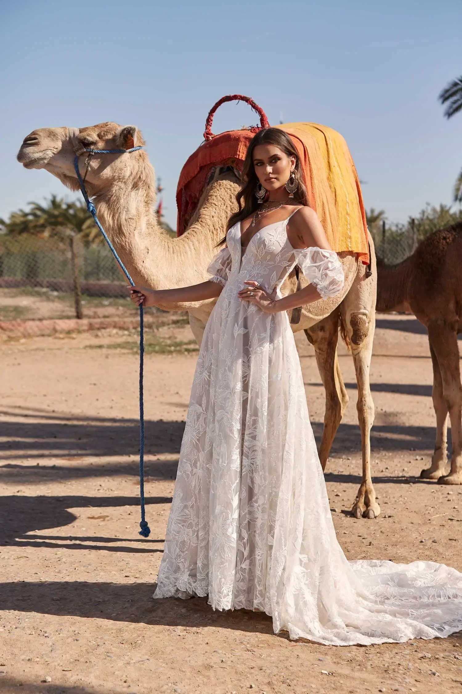 A woman in a flowing white lace gown stands confidently next to a camel draped in orange fabric. The scene is bright, evoking a sense of elegance and adventure.