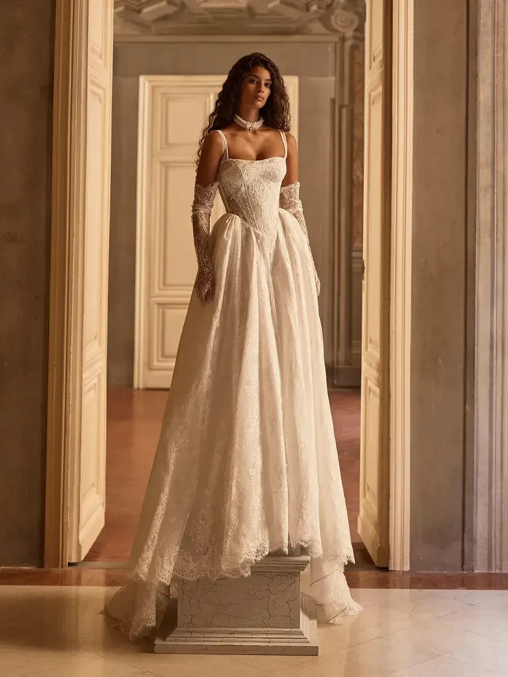 A model wearing a long, elegant white gown, standing on a pedestal in a beautifully ornate interior.