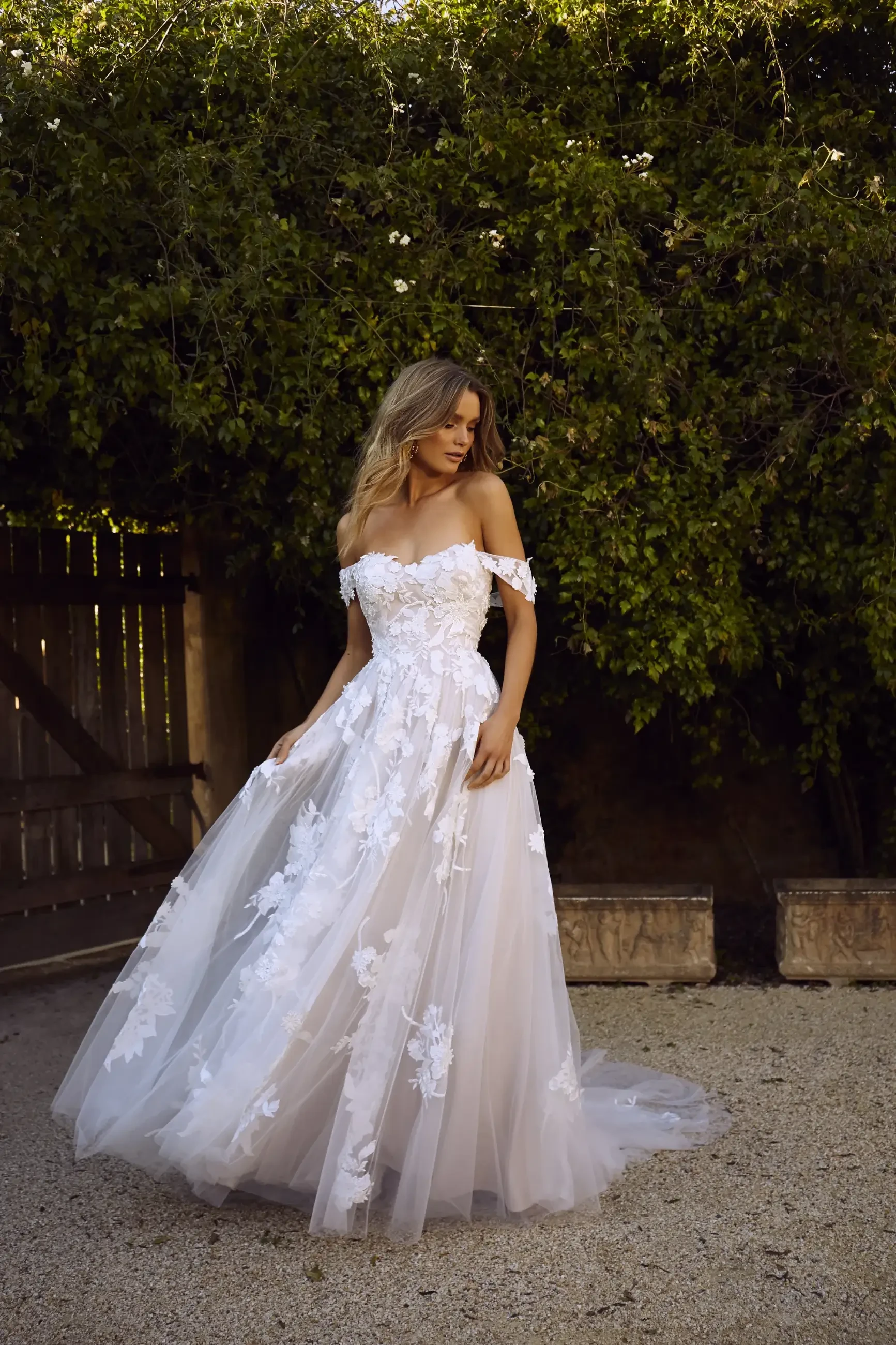 A bride stands in an outdoor setting wearing an off-the-shoulder, white floral lace wedding gown. She gazes downwards, surrounded by lush greenery.