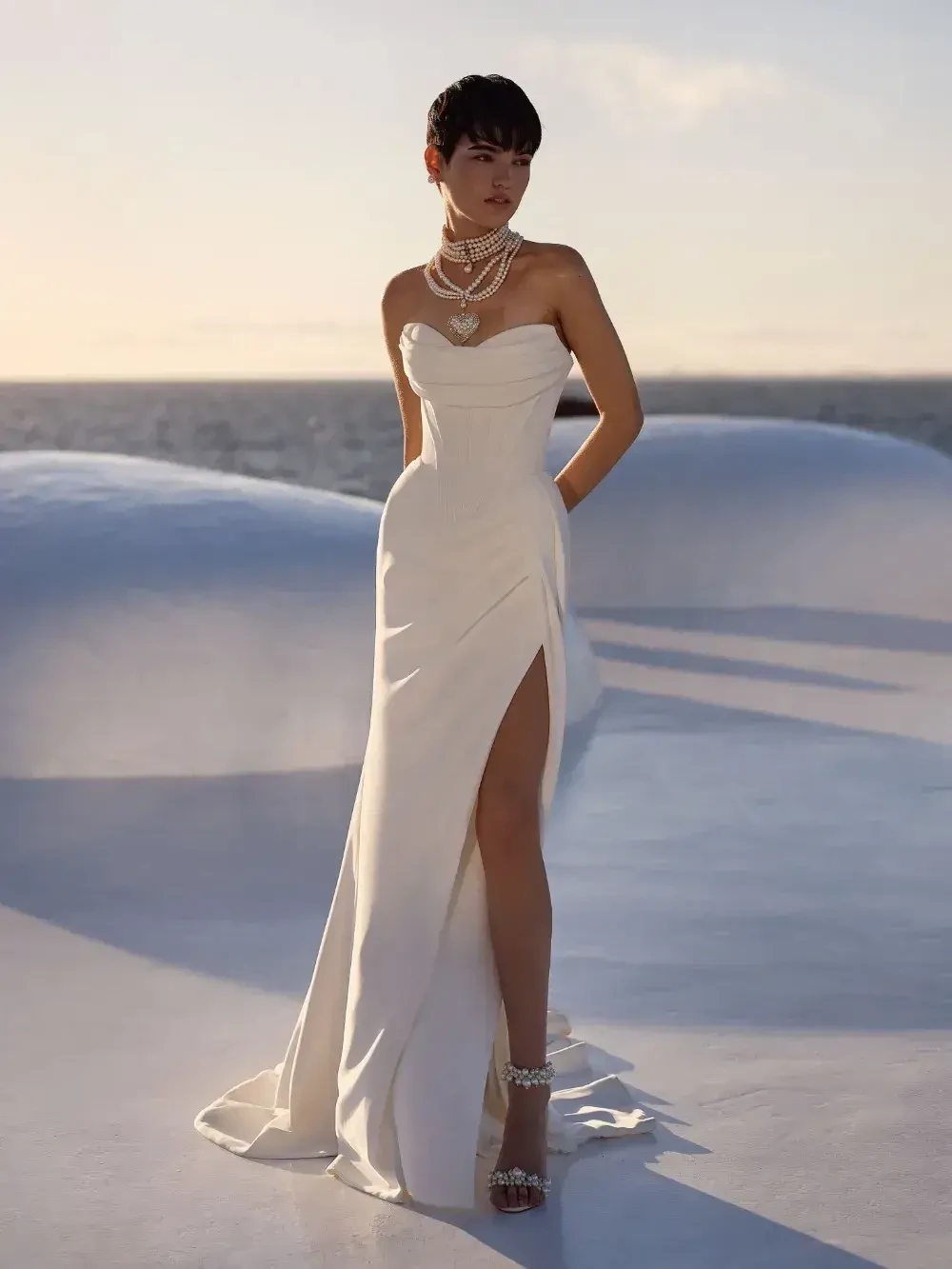 A woman wearing a strapless white gown with a thigh-high slit, styled with layered pearl necklaces, standing on a sunny outdoor setting near the sea.