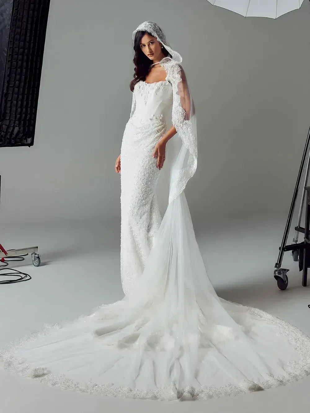 A model wearing a white wedding dress with a long train and a hood, standing in a studio setting.