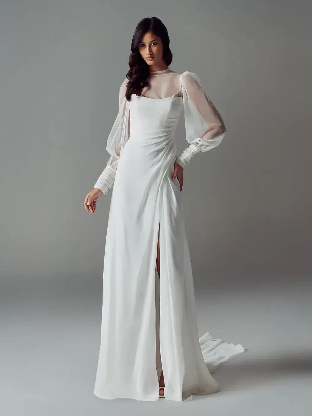 A woman wearing a white long-sleeve gown with a high slit and sheer sleeves, posed against a simple gray background.