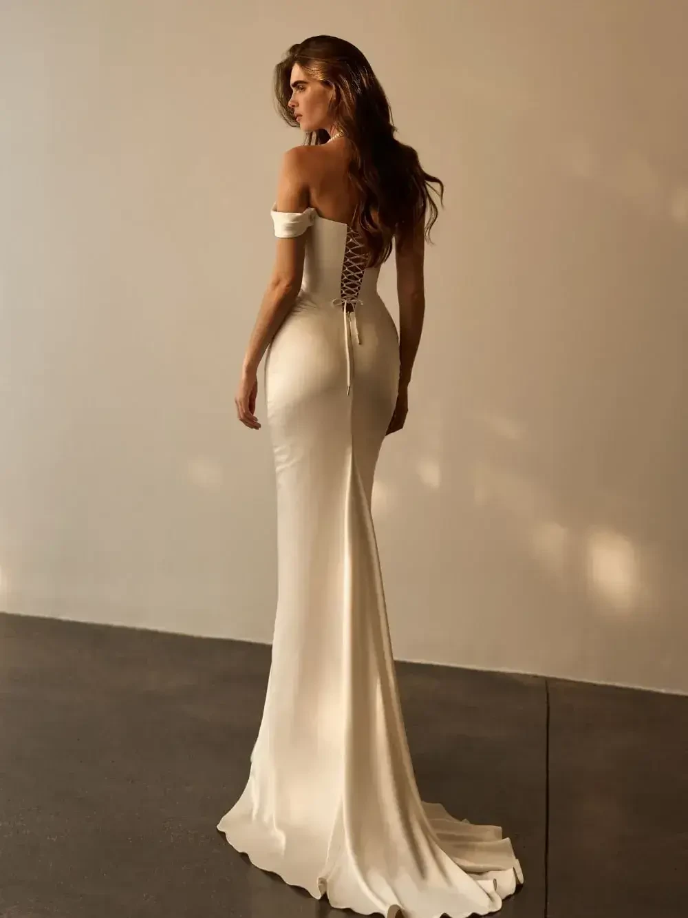 A woman in a fitted white dress with off-the-shoulder straps and a lace-up back, standing with her back facing the camera.