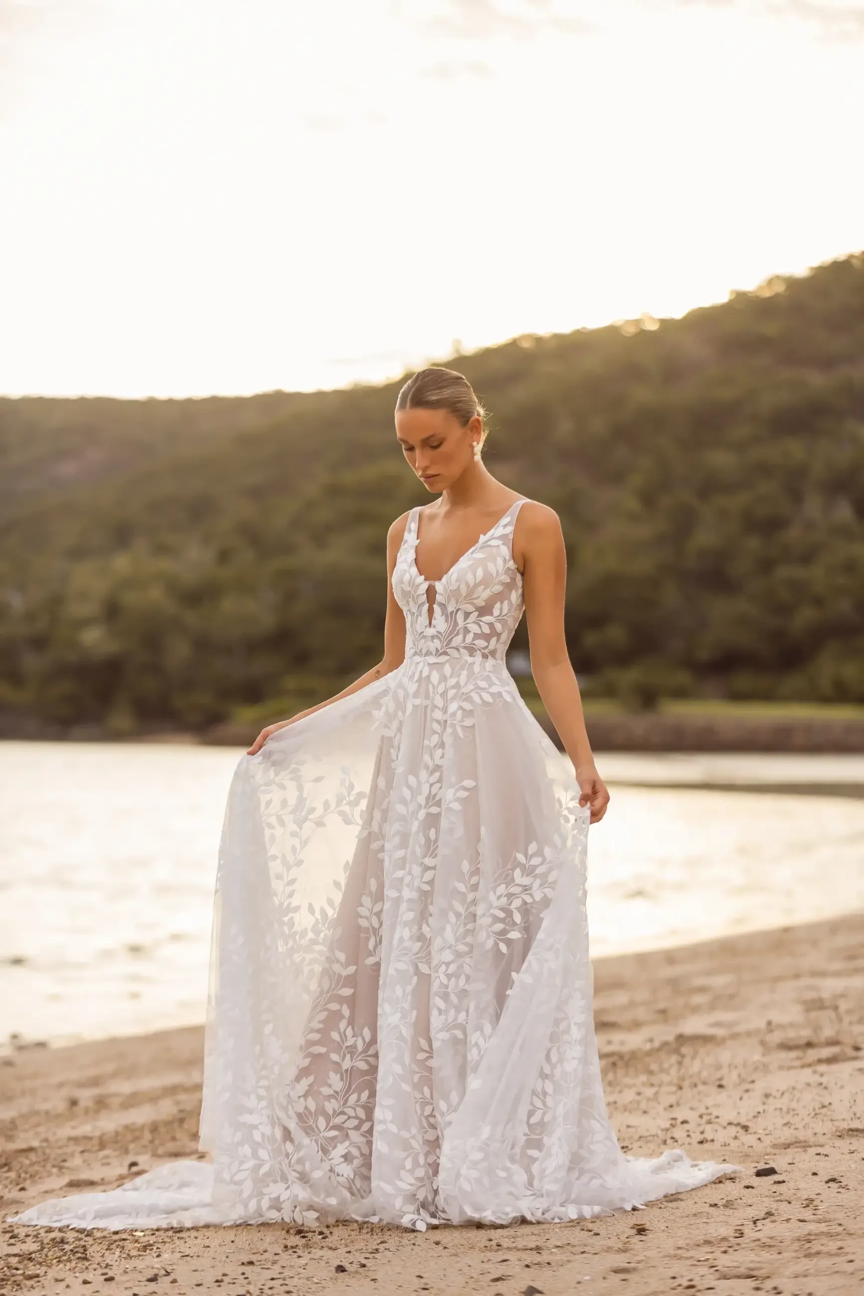 A woman in a flowing white dress with floral lace detailing stands on a beach near the water during sunset.