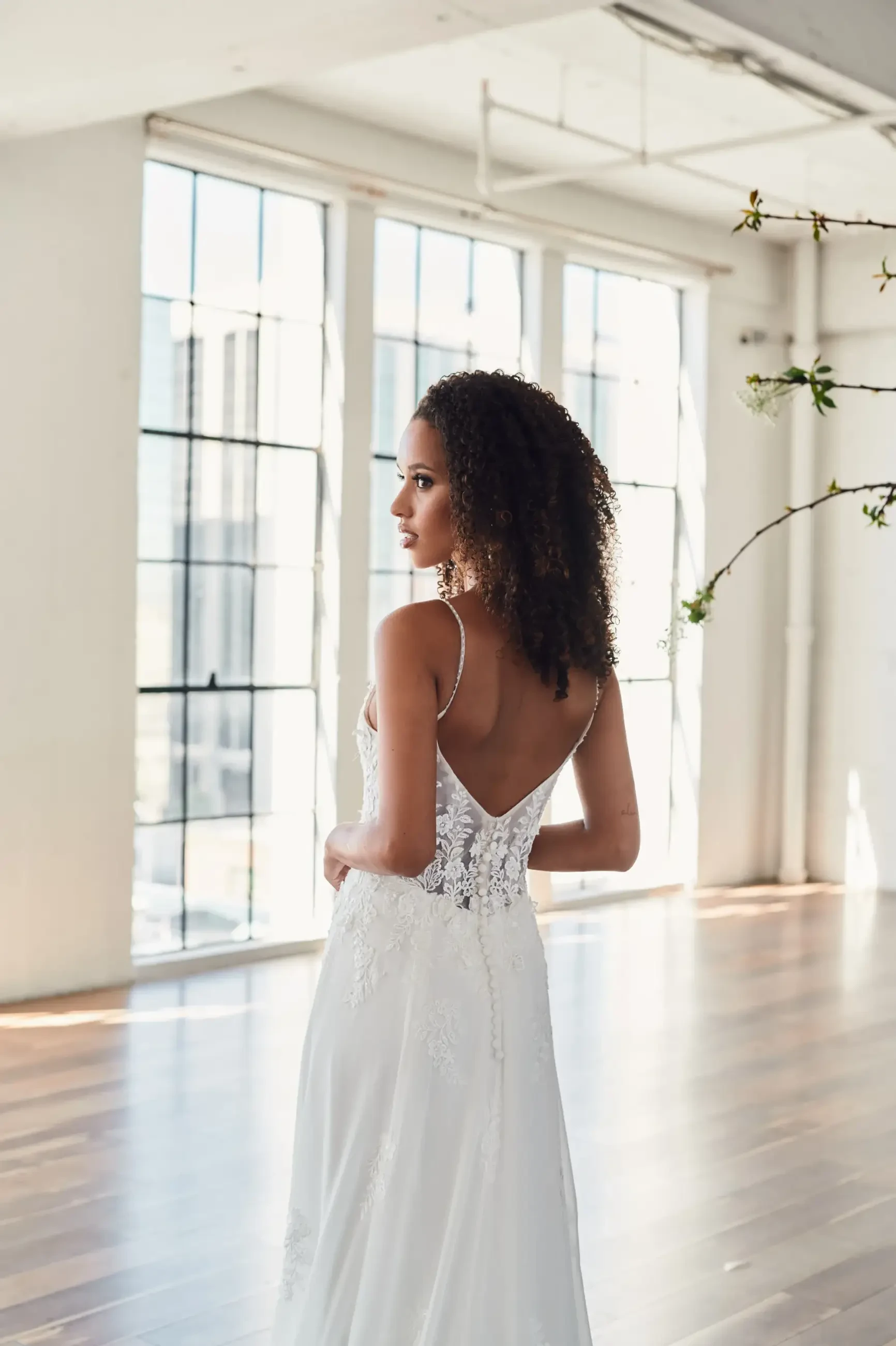 Bride in a lace wedding dress with spaghetti straps stands in a sunlit loft, gazing out tall windows. The atmosphere is airy and serene.