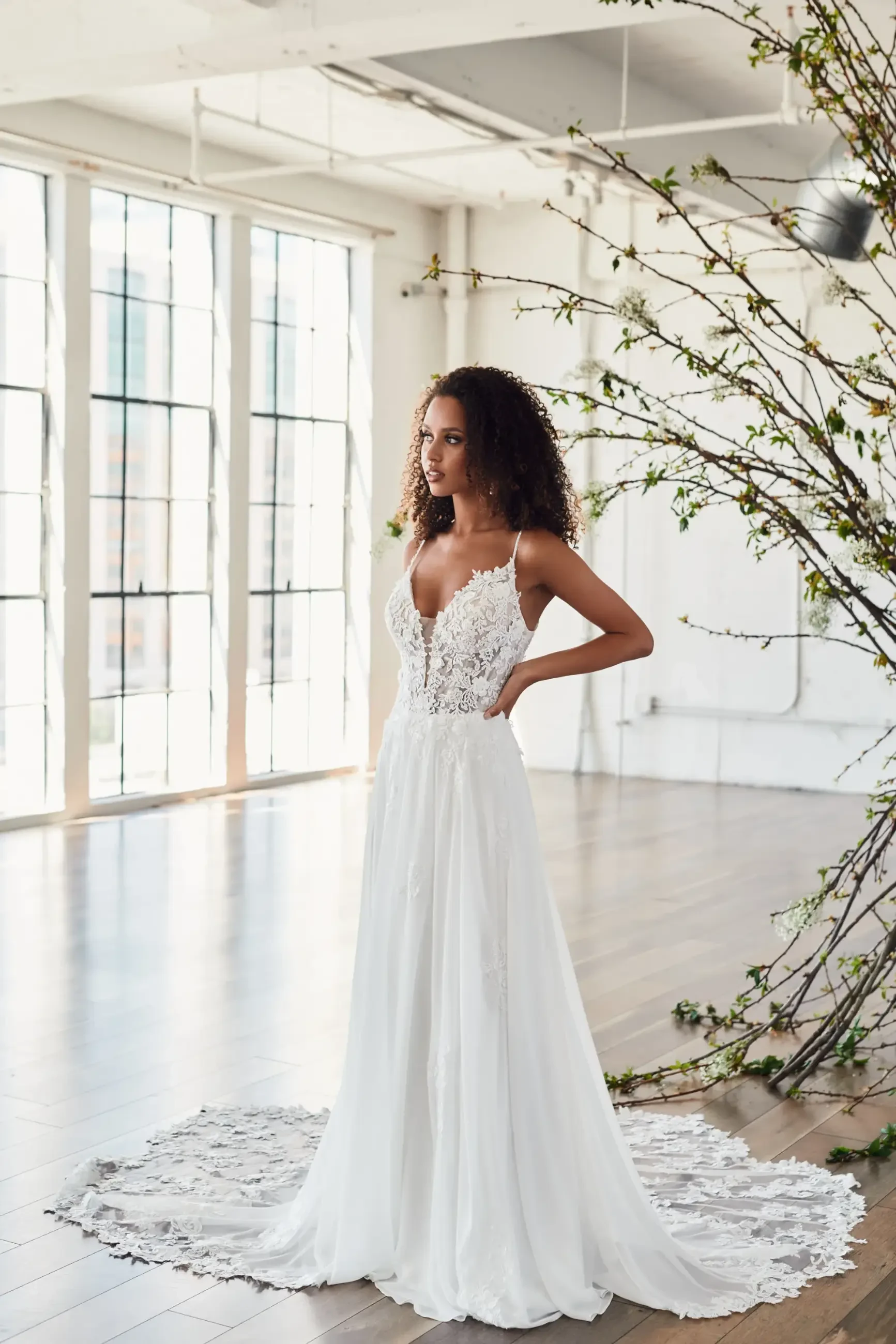 A woman in a flowing white lace wedding dress stands in a bright room with large windows. Branches with green leaves add natural elegance to the scene.