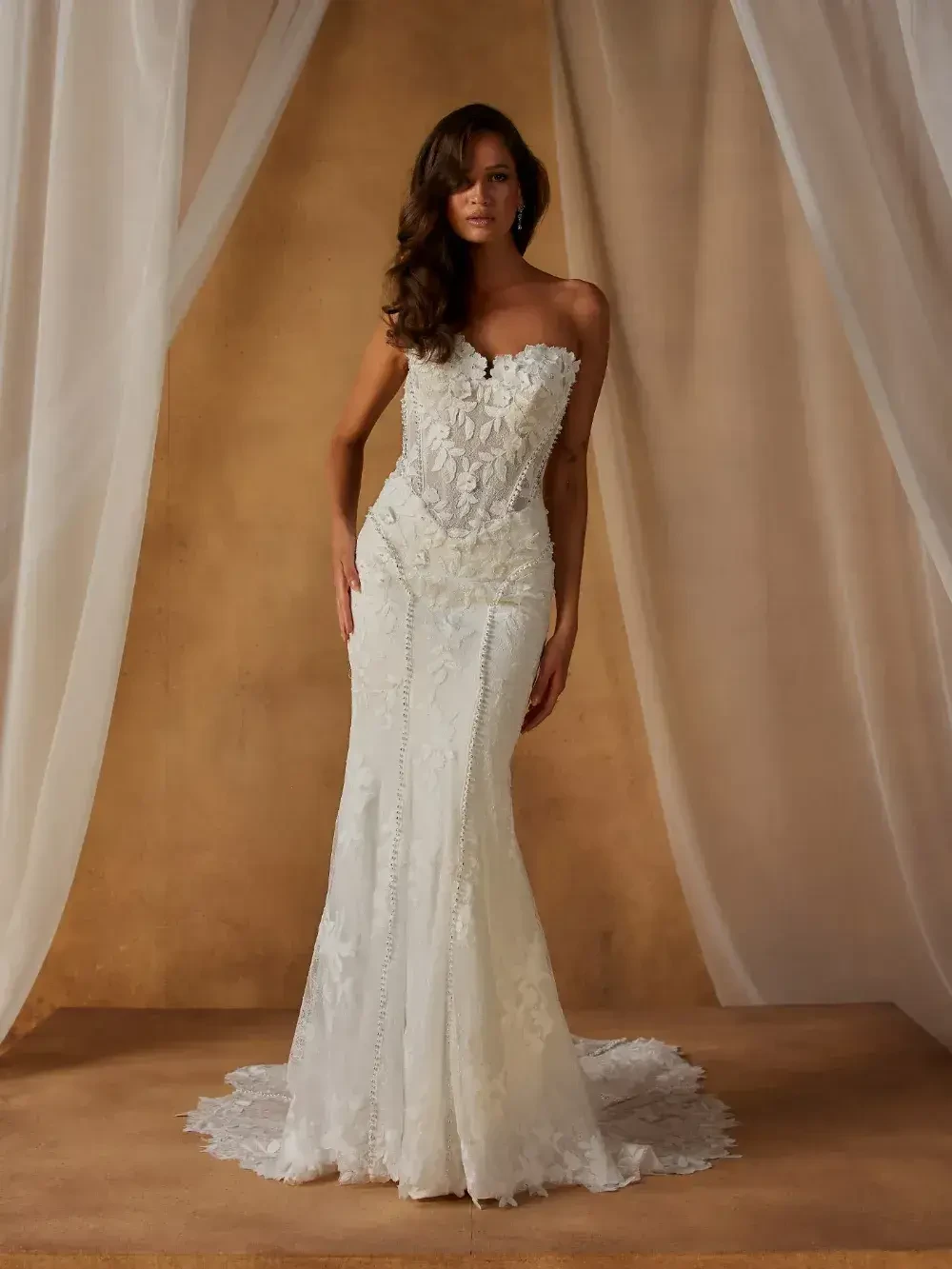 A woman wearing a strapless white wedding dress with intricate lace details, posing against a draped background.