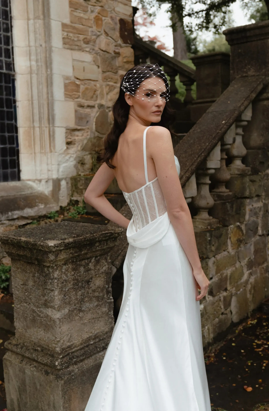 Bride in elegant white gown with sheer back and pearl straps stands by a stone staircase, wearing a retro netted headpiece, conveying a vintage feel.