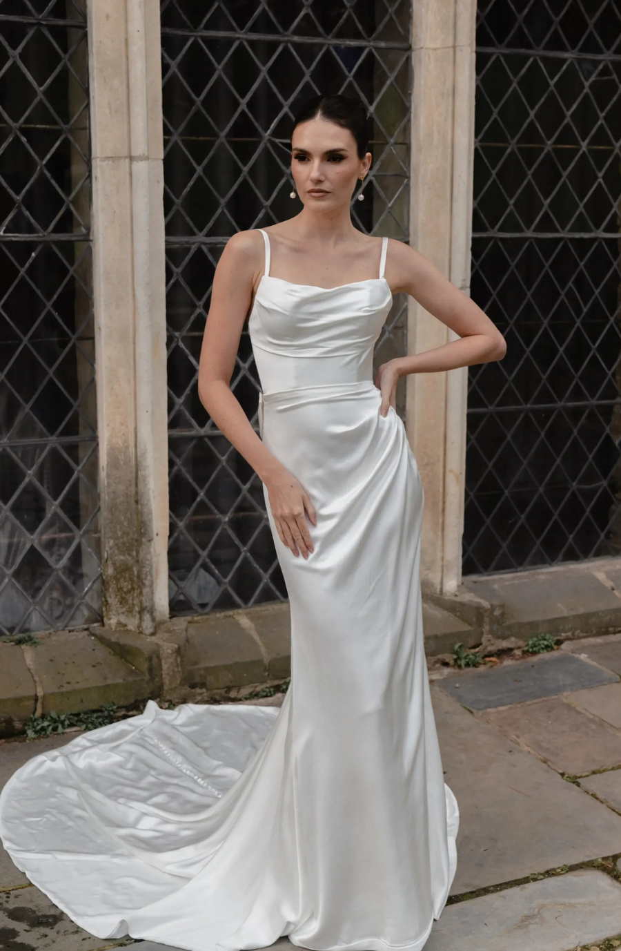 A woman in an elegant white satin gown stands confidently, hand on hip, against a backdrop of stone and diamond-patterned windows, conveying sophistication.