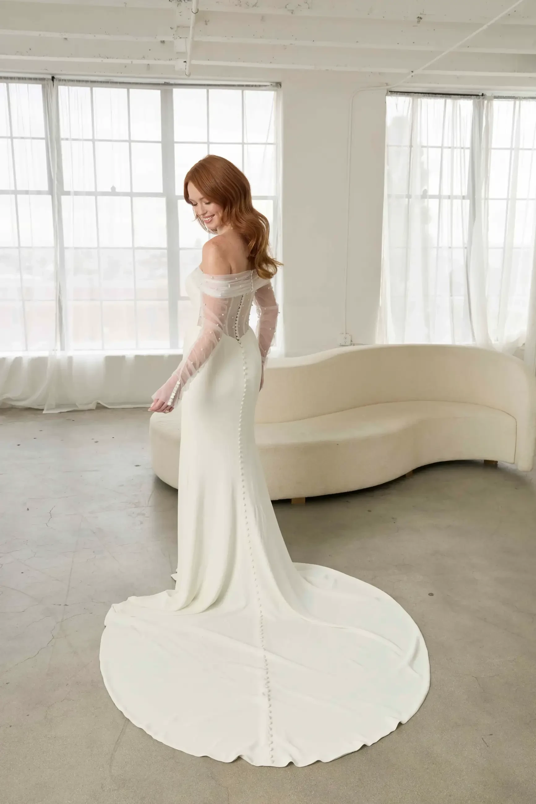 A woman in a white wedding dress poses with her back to the camera, showcasing the train of the gown in a well-lit room.