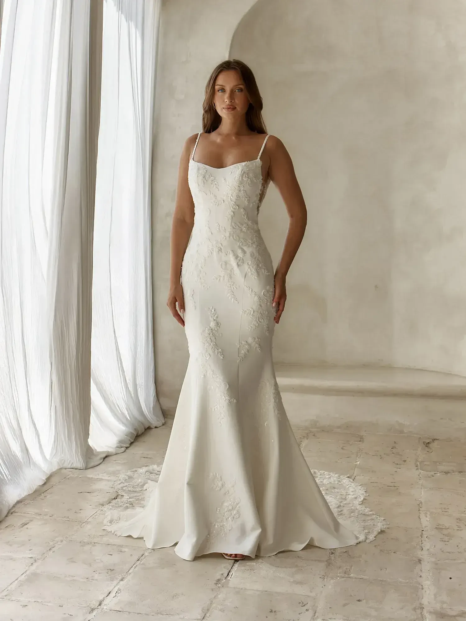 A woman stands in a white wedding dress with spaghetti straps, featuring intricate embroidery, against a neutral backdrop.