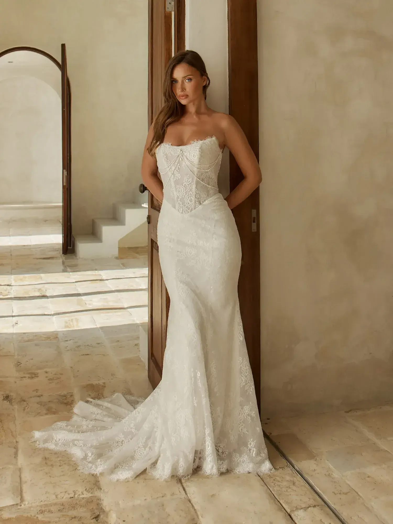 A woman in a strapless white wedding dress stands elegantly next to a wooden door in a spacious, softly lit room with stone flooring.