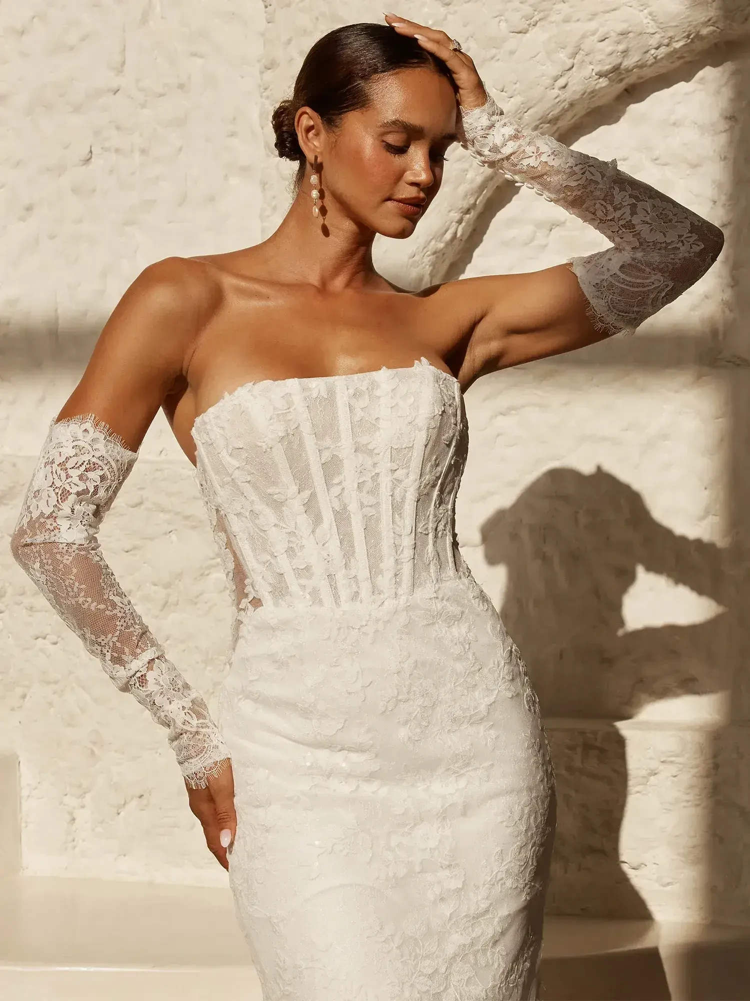 A model in a strapless white lace wedding dress with sheer lace arm gloves, posing against a textured white wall.